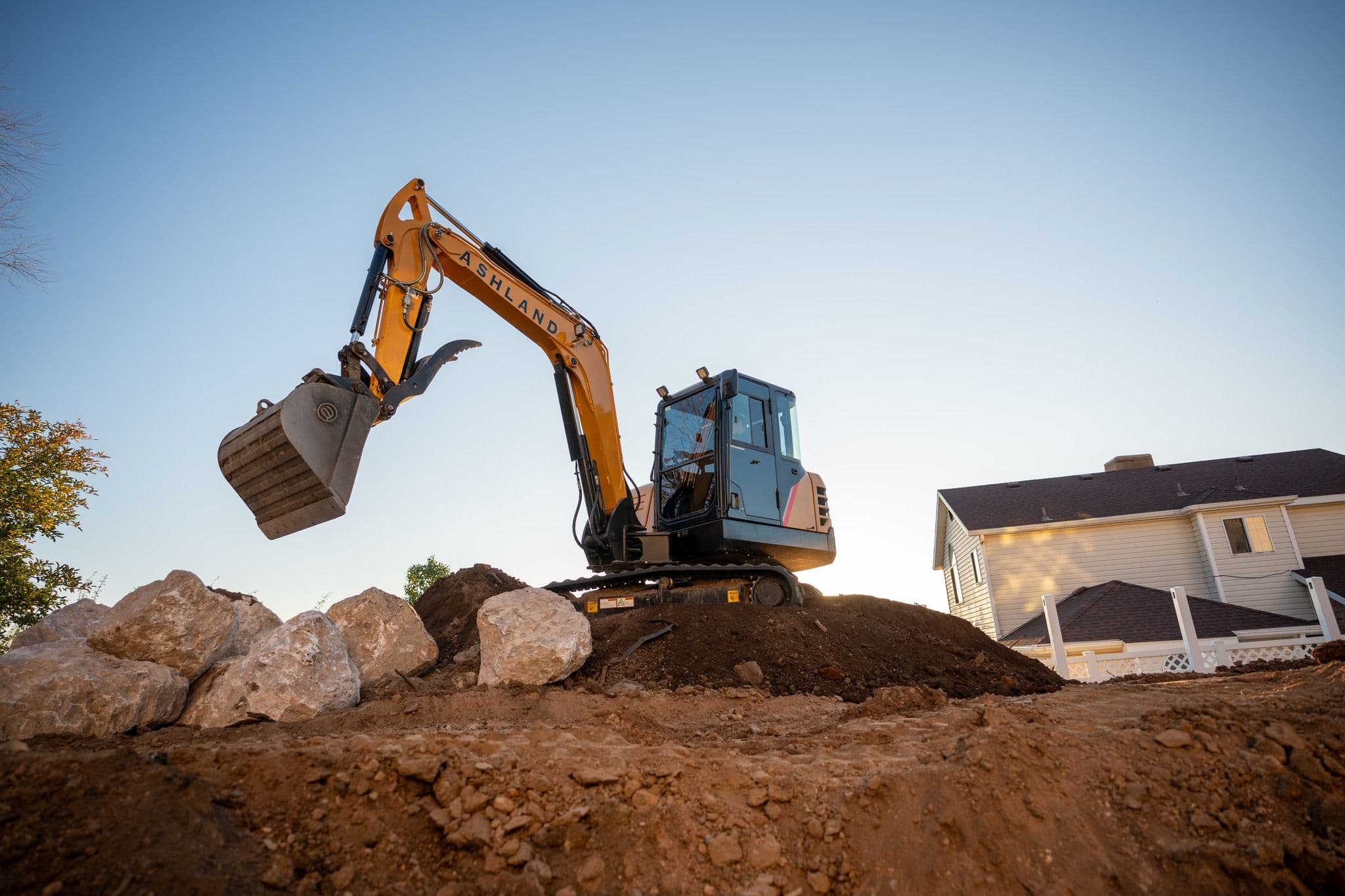 Excavator digger on a construction site, moving dirt and rocks near a house.