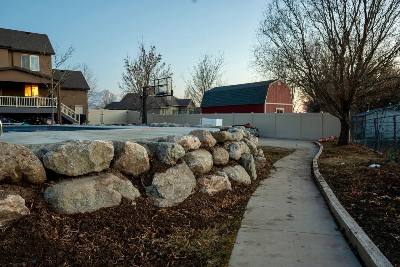 Residential backyard with stone retaining wall, sidewalk, and barn under a sunset sky.